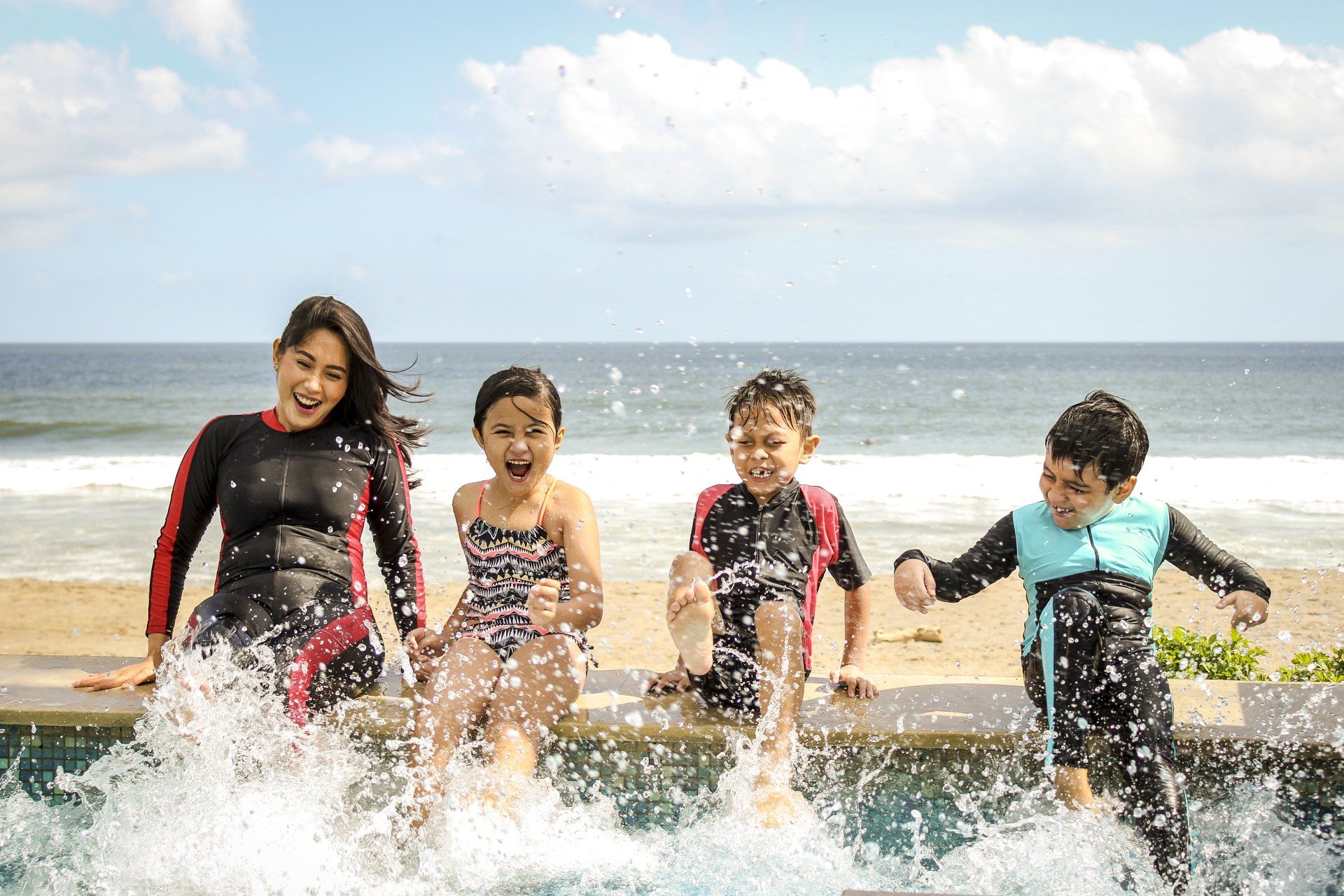 Four children in wetsuits splashing and laughing in shallow ocean waves on a sunny beach.